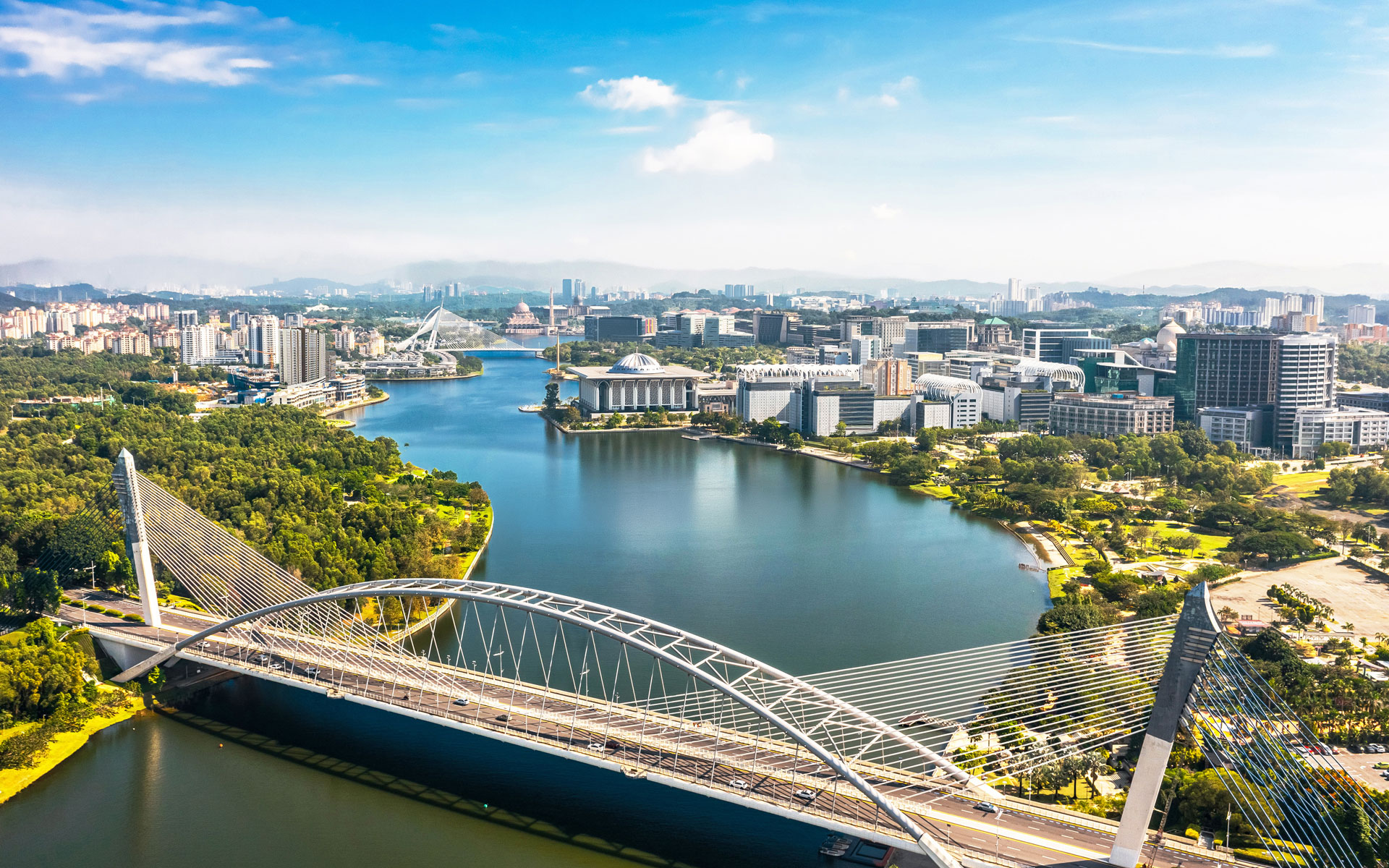 Aerial view of Putrajaya featuring its iconic curved bridge over a blue lake, modern government buildings, lush greenery, and scenic skyline
